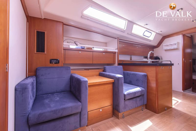 De foto toont het interieur van een boot met twee blauwe fauteuils, een houten tafel en een keukenblok met spoelbak.