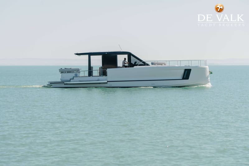 Een witte motorboot vaart op helderblauw water, met een persoon aan het roer en een logo van "DE VALK YACHT BROKERS"…