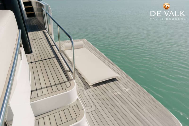 Een houten dek met een ligbed en een reling, met op de achtergrond water en het logo van "DE VALK YACHT BROKERS".
