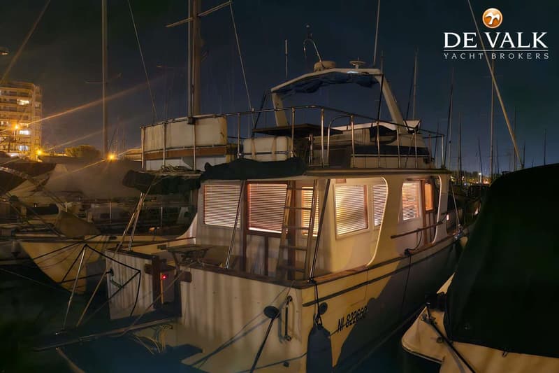 Een verlichte boot met ramen met jaloezieën ligt 's nachts in een haven, met andere boten en gebouwen op de achtergrond.