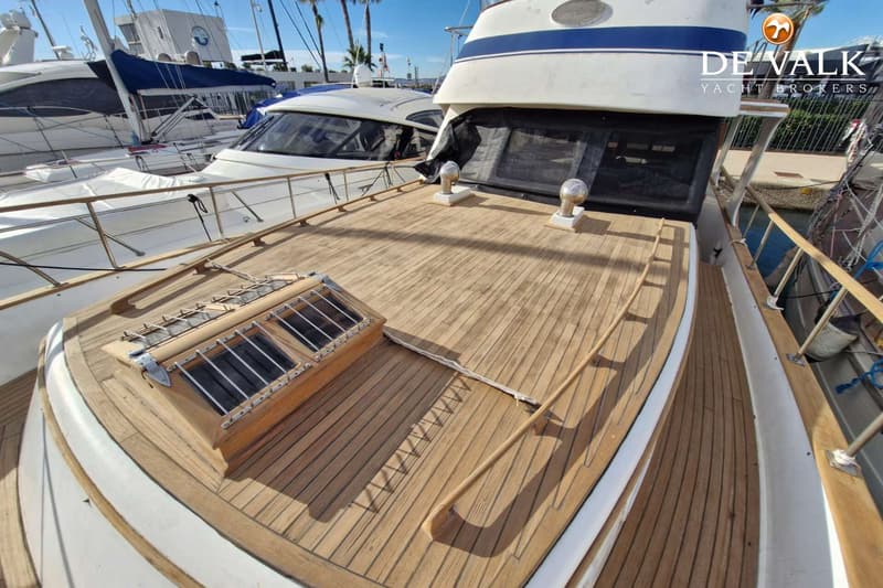 Een houten dek met een luik en twee ventilatiekappen, met een deel van een witte boot en een blauwe streep zichtbaar.