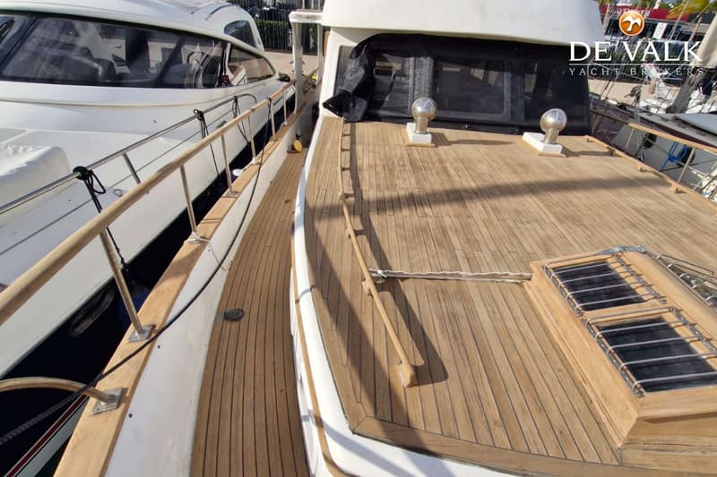 Een bovenaanzicht van het teakhouten dek van een boot met een luik en een reling, naast een andere boot.
