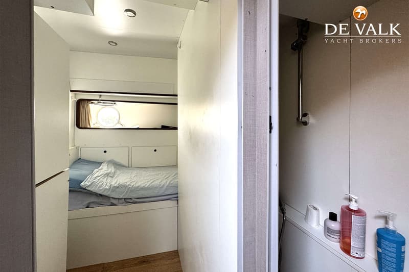 Een kajuit met een bed met blauw beddengoed en een patrijspoort, en een deels zichtbare doucheruimte met flessen zeep.