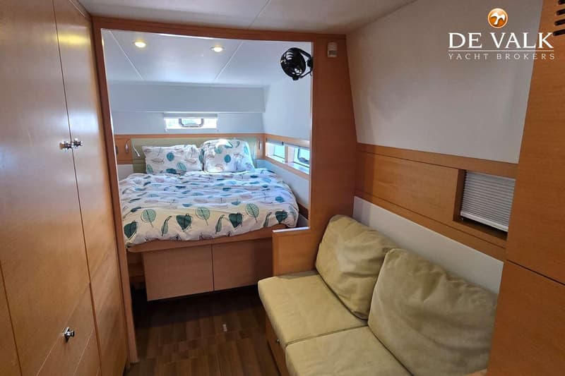 Een kajuit met een bed met bladmotief beddengoed, een bank met kussens, houten kasten en een ventilator. — Nautitech 542