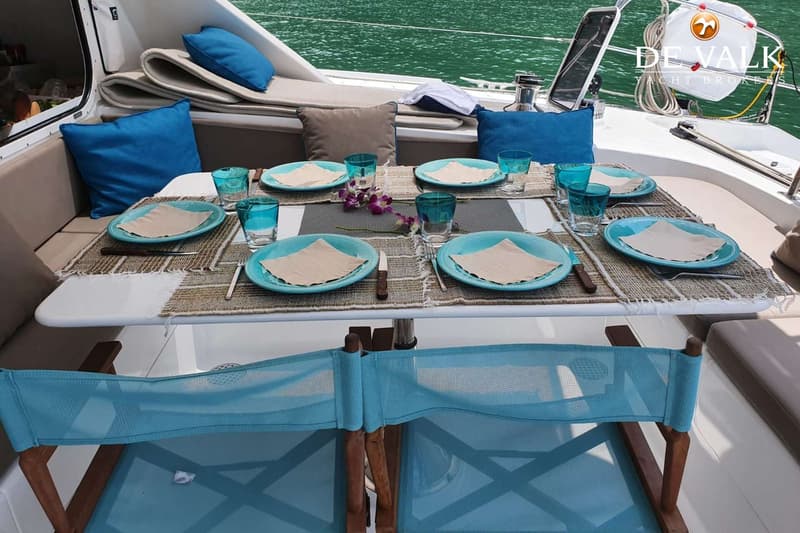 Een gedekte tafel op een boot met blauwe borden, glazen, bestek en servetten, omringd door stoelen en kussens.