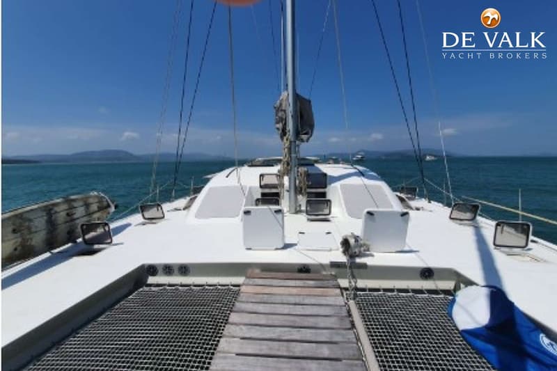Een witte catamaran met een mast en zeilhoes, met een houten loopplank en netten aan de voorkant, vaart op zee.