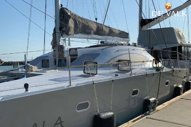 Een grijze catamaran met een opgerold zeil ligt aangemeerd aan een houten steiger onder een helderblauwe lucht.