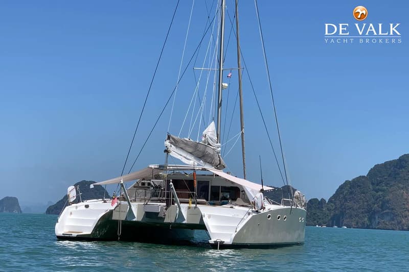 Een witte catamaran met twee masten en opgerolde zeilen ligt op helderblauw water met groene, rotsachtige eilanden op de…