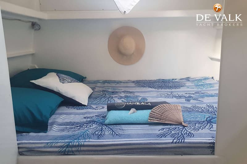Een kajuit met een bed met blauw-wit gestreept en koraalpatroon beddengoed, twee kussens, een hoed en handdoeken.