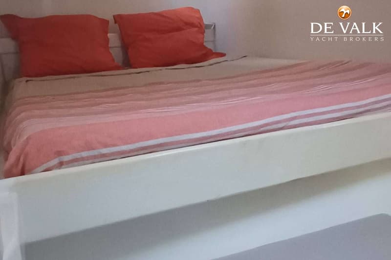 Een bed met roze gestreepte dekens en twee oranje kussens is zichtbaar, met een logo van "DE VALK YACHT BROKERS".