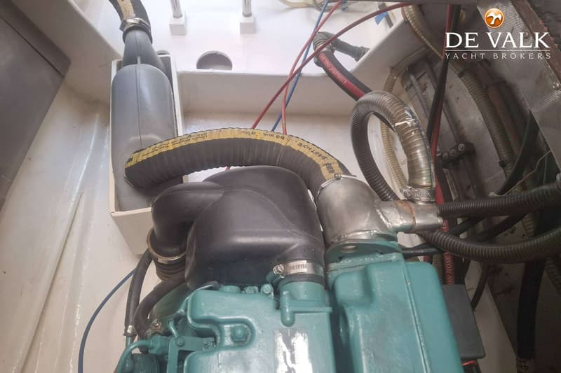 Een close-up van een motorruimte met een turquoise motorblok, diverse slangen en kabels, en het logo van "DE VALK YACHT…