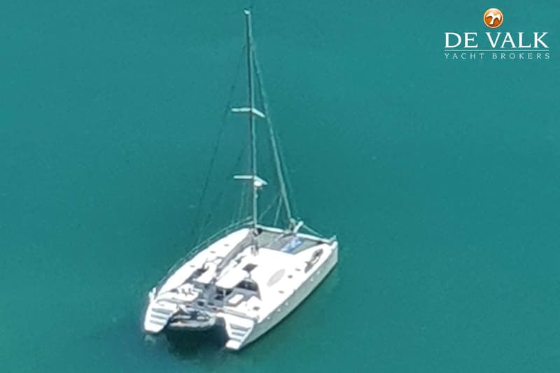 Een witte catamaran met een mast en tuigage ligt in turquoise water, gezien van bovenaf.