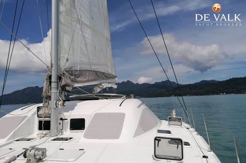 Een witte catamaran met opgetrokken zeilen vaart op helderblauw water, met bergen op de achtergrond onder een bewolkte lucht.