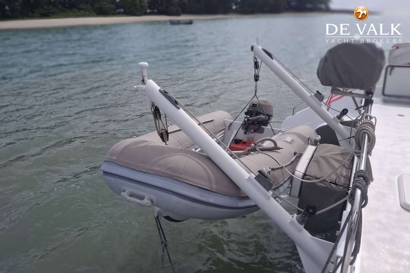 Een grijze bijboot met buitenboordmotor hangt aan davits boven het water, met een zandstrand en bomen op de achtergrond.