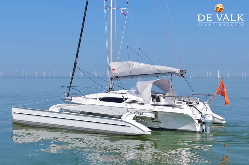 Een witte trimaran met een Nederlandse vlag en een Honda buitenboordmotor, varend op rustig water met windmolens op de…