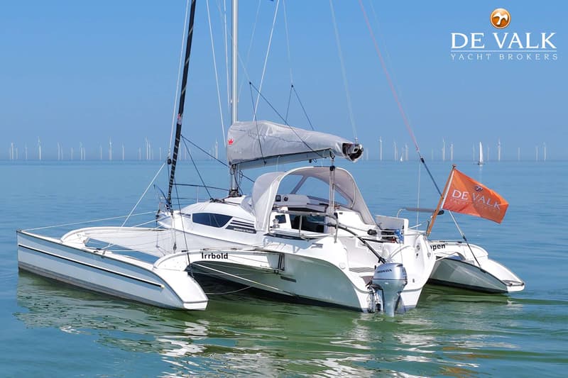 Een witte catamaran met een buitenboordmotor en een oranje vlag vaart op rustig water met windmolens op de achtergrond.
