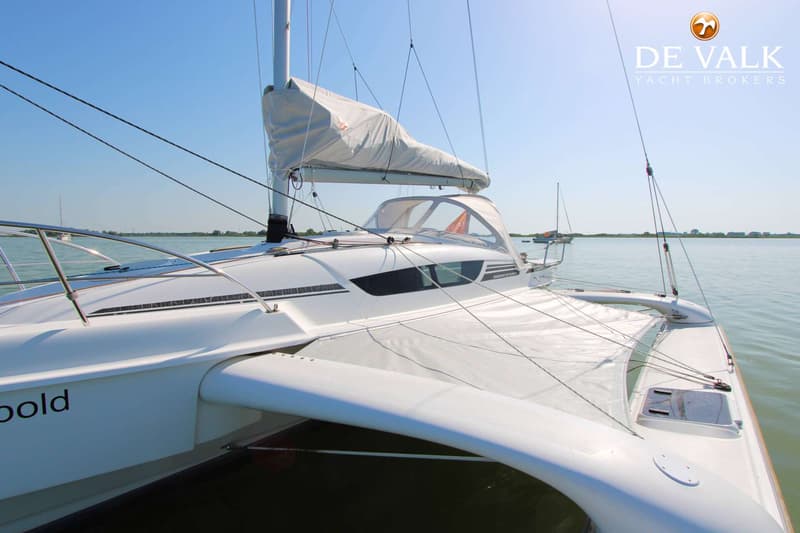 Een witte trimaran met een opgerold zeil en een kajuit met ramen, drijvend op rustig water onder een helderblauwe lucht.