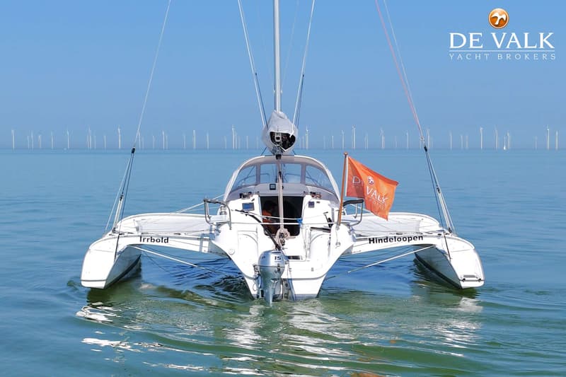 Een witte catamaran met een buitenboordmotor en een oranje vlag vaart op rustig water met windmolens op de horizon.