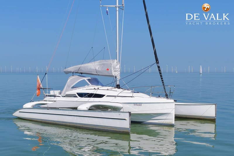 Een witte trimaran met de naam "Irrbold" ligt op rustig water, met windmolens aan de horizon.
