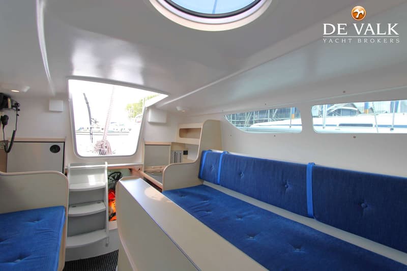 De foto toont het interieur van een boot met blauwe kussens op banken, een patrijspoort en een luik in het plafond.