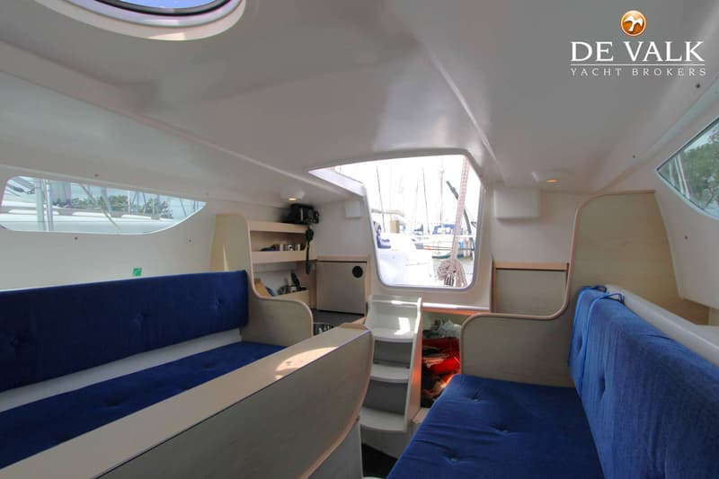 De foto toont het interieur van een boot met blauwe zitkussens, een kajuitingang en ramen met uitzicht op andere boten.