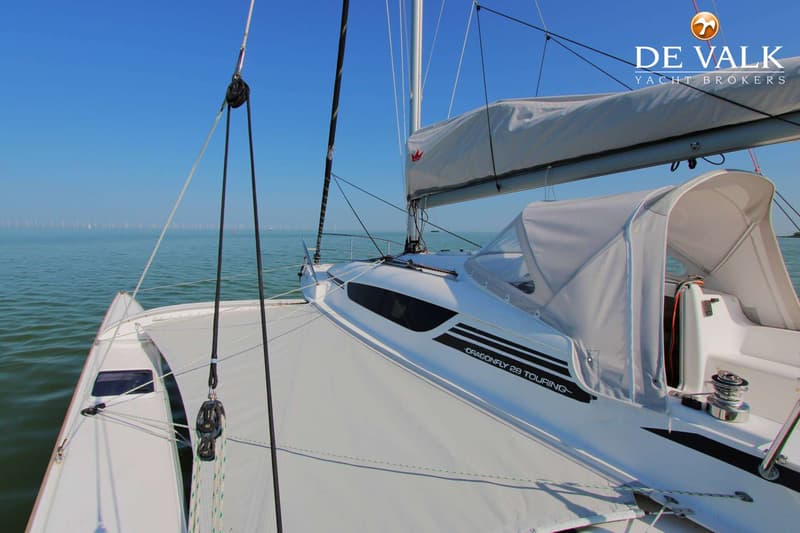 Een witte trimaran met de tekst "DRAGONFLY 28 TOURING" op de romp, vaart op kalm water onder een helderblauwe lucht.