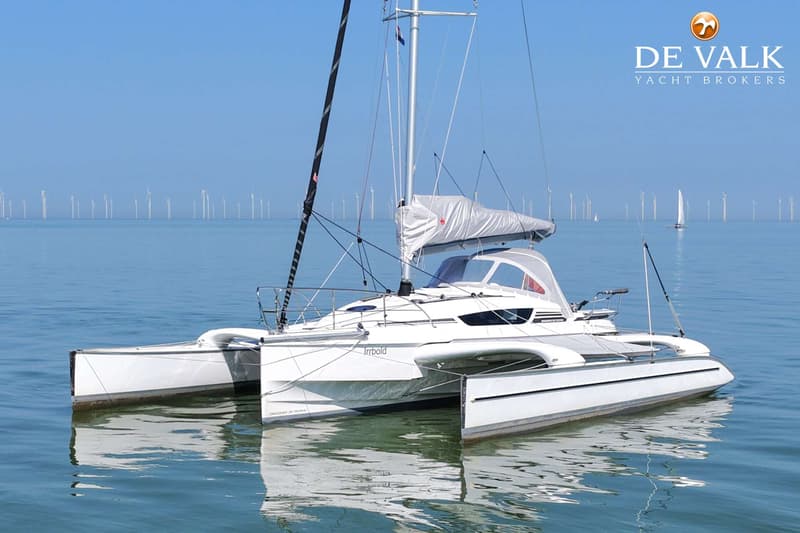Een witte trimaran met de naam "Irrbold" ligt op rustig water met windmolens op de achtergrond.