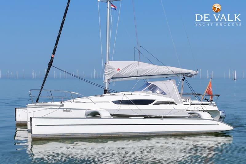 Een witte trimaran met de naam "Irrbold" ligt op het water met een grijze zeilhoes en een Nederlandse vlag.