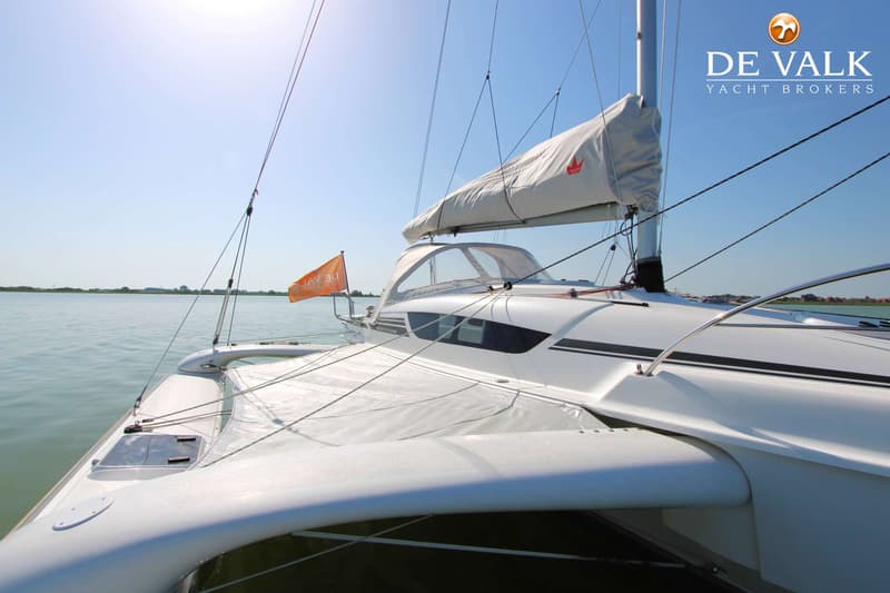 Een witte catamaran met een opgerold zeil en een oranje vlag vaart op kalm water onder een heldere hemel.