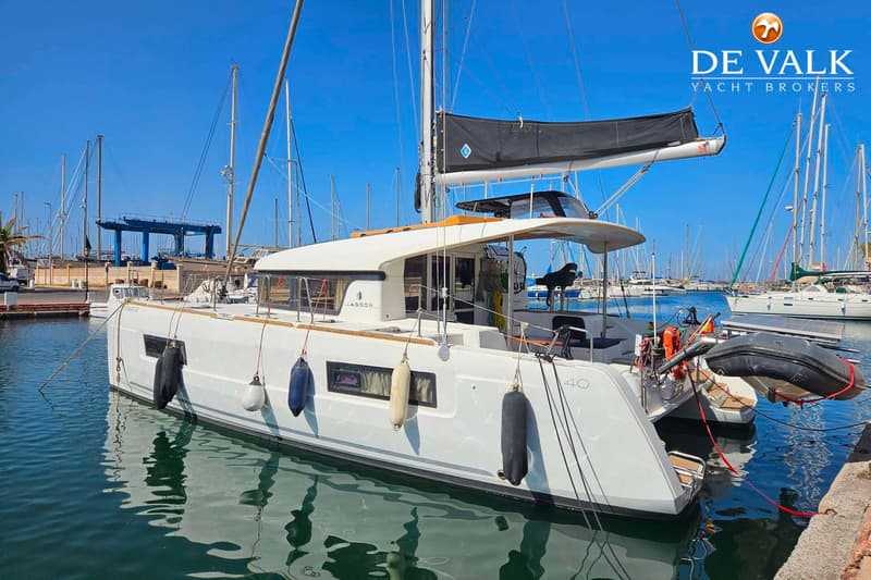 Een witte catamaran met een zwart zeil ligt afgemeerd in een haven, met andere boten op de achtergrond.