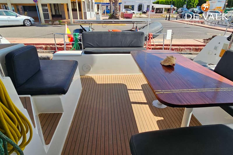 Een bootdek met teakhouten vloer, een houten tafel met een schelp erop, en zwarte kussens op de zitbanken.