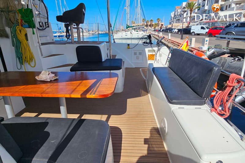 Een houten tafel met een schelp erop, omringd door zitplaatsen met zwarte kussens, op een bootdek met uitzicht op een haven.
