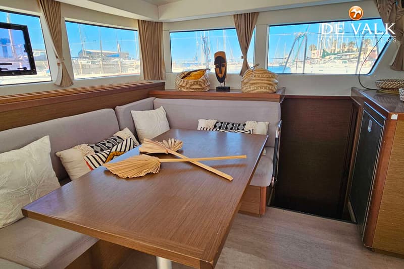 Een interieur van een boot met een houten tafel, grijze banken met kussens en ramen met uitzicht op een haven. — Lagoon 40