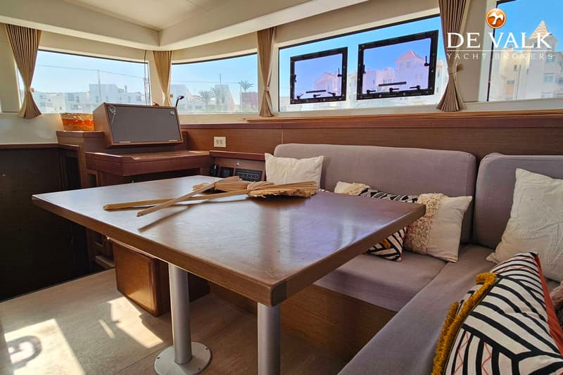 Een interieur van een boot met een houten tafel, een grijze bank met kussens en ramen met uitzicht op gebouwen. — Lagoon 40