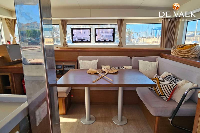 Een interieur van een boot met een houten tafel, grijze banken met kussens, en ramen met uitzicht op een haven. — Lagoon 40