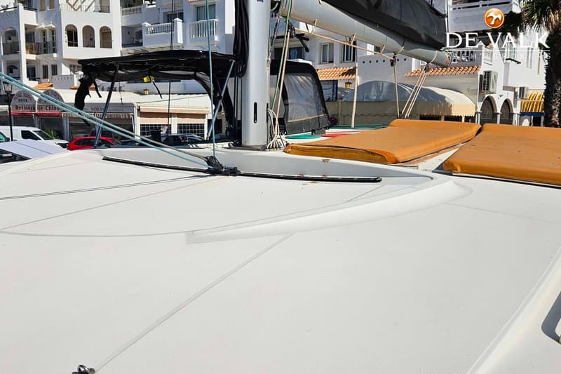Een wit dek van een boot met een mast, zeil, touwen en oranje kussens, met gebouwen op de achtergrond. — Lagoon 40