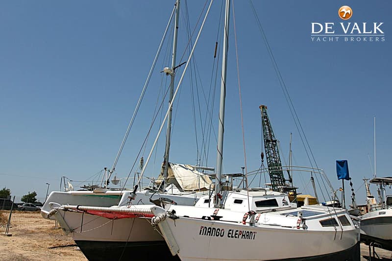 Twee witte catamarans met masten en tuigage staan op het droge, met een kraan en andere boten op de achtergrond.