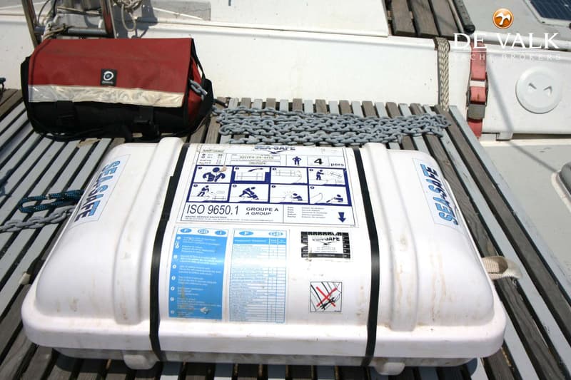 Een witte reddingsvlotcontainer van het merk "SEA-SAFE" ligt op een houten roosterdek, met een rode tas en ankerketting…