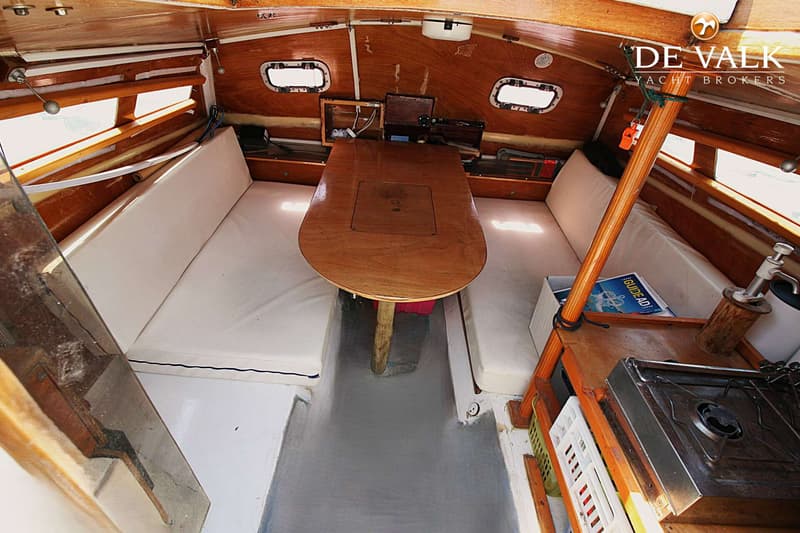 De foto toont het interieur van een boot met houten wanden, een tafel, zitbanken en een kooktoestel. — Wharram Tiki 30