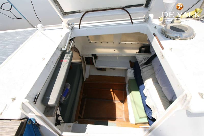Een open kajuitingang met een houten vloer, een bed met kussens en een Raymarine stuurautomaat.