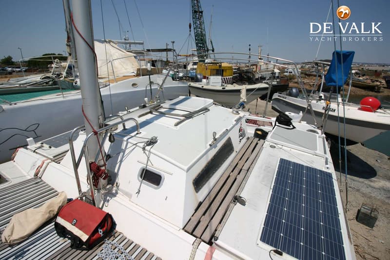 Een witte zeilboot met zonnepanelen op het dek, liggend in een haven met andere boten en een kraan op de achtergrond.