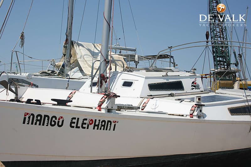 Een witte zeilboot met de naam "MANGO ELEPHANT" op de zijkant, met een kraan en andere boten op de achtergrond.
