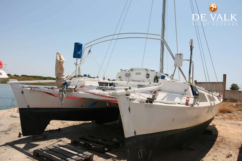 Een witte catamaran met zwarte onderkant staat op het droge, met een blauwe lucht en water op de achtergrond.