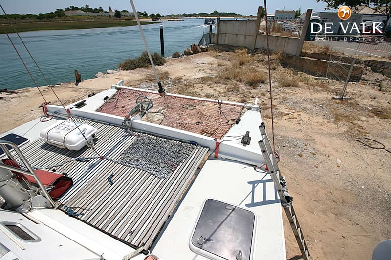 Een bovenaanzicht van de boeg van een catamaran met een reddingsvlot, ankerketting en netten, liggend aan een kade.