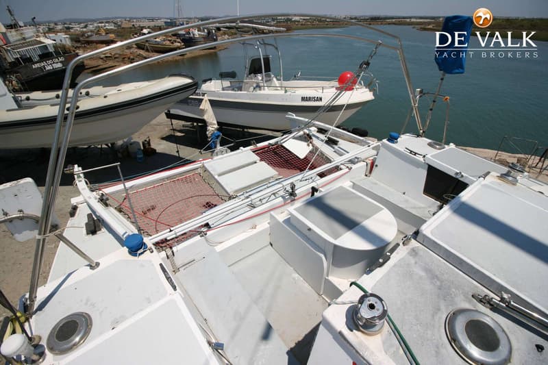 Een bovenaanzicht van het dek van een witte catamaran met een rood net, liggend aan de wal naast andere boten.