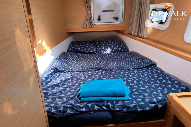 Een kajuit met een tweepersoonsbed met donkerblauw beddengoed met een patroon, twee kussens en een opgevouwen blauwe…