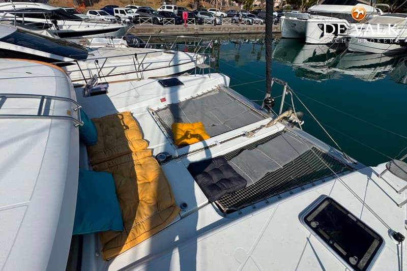 Een bovenaanzicht van het voordek van een catamaran met ligkussens en netten, afgemeerd in een haven.