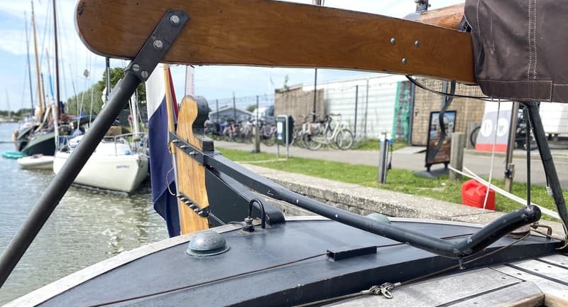 Een houten giek met metalen beslag en een Nederlandse vlag op de achtergrond, met andere boten in het water.