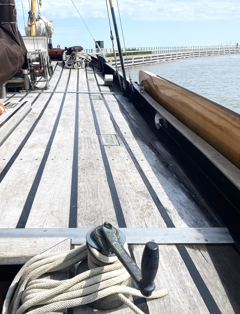 Een houten dek met donkere naden, een lier met touw, en een deel van de romp van een boot zijn zichtbaar.