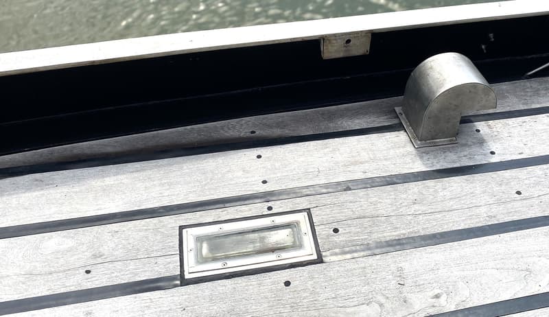 Een houten dek met zwarte naden, een metalen ventilatiekap en een rechthoekig glazen luik.